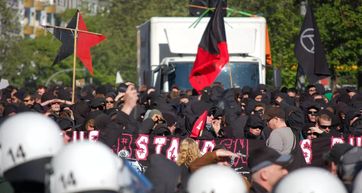 Im Vordergrund Polizeihelme, dahinter schwarz gekleidete Demonstrant:innen mit schwarz-roten Fahnen.