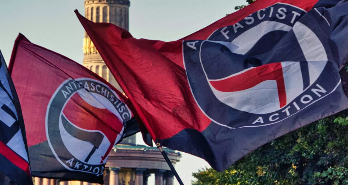 Fahnen mit dem Antifa-Logo, im Hintergrund die Berliner Siegessäule. Das Antifa-Logo besteht aus einer roten und einer schwarzen Flagge in einem Kreis sowie dem Schriftzug "Antifaschistische Aktion".